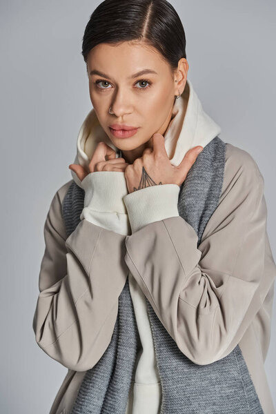 A young woman exudes style in a gray coat and white turtle neck sweater against a studios gray backdrop.