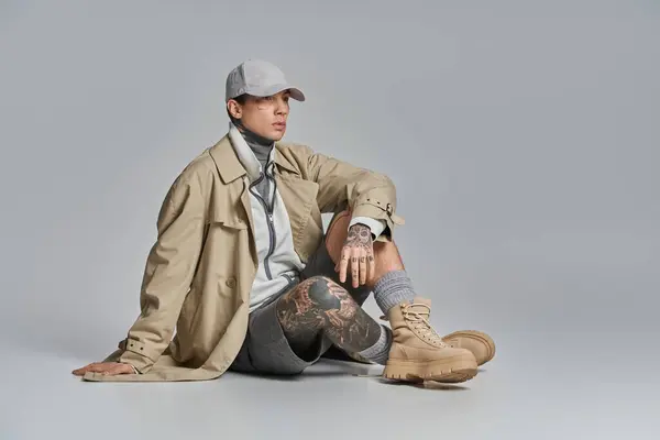一个身穿战壕外套 纹身的年轻人坐在地上 两条腿交叉在一起 陷入沉思 与灰色的工作室背景相映衬 — 图库照片