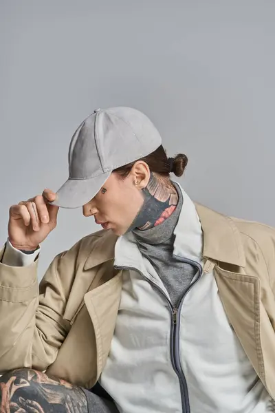 stock image A young man with a tattoo on his face sits, wearing a trench coat, in a studio against a grey background.