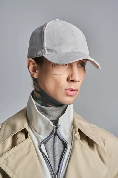 一个年轻的纹身男子穿着时髦的风衣 头戴时髦的帽子 在灰色的工作室背景下散发出一股清凉的气息 — 图库照片