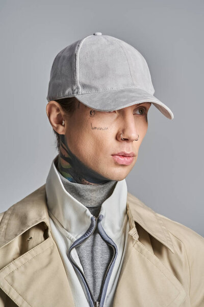 Молодой, татуированный мужчина излучает прохладу в стильном плаще, в комплекте с модной шляпой, на сером фоне студии.
