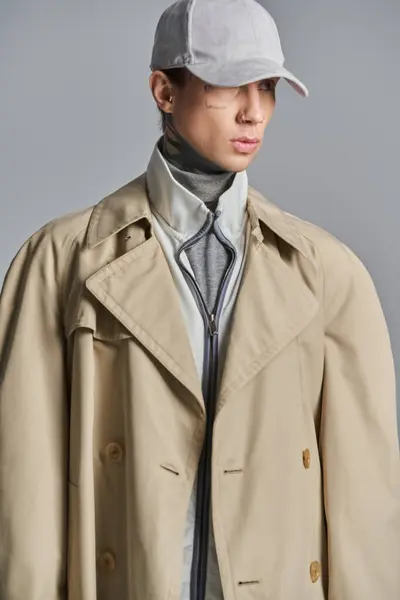 stock image A young, tattooed man exudes mystery and style in a trench coat and hat, set against a grey studio backdrop.