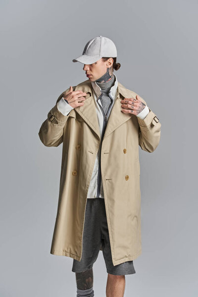 A young, tattooed man stands in a trench coat and hat, exuding mystery and intrigue.