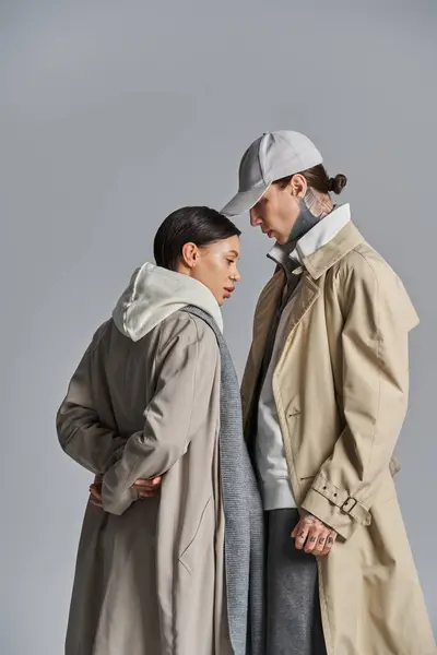 stock image A young stylish couple in trench coats standing together in a studio on a grey background.