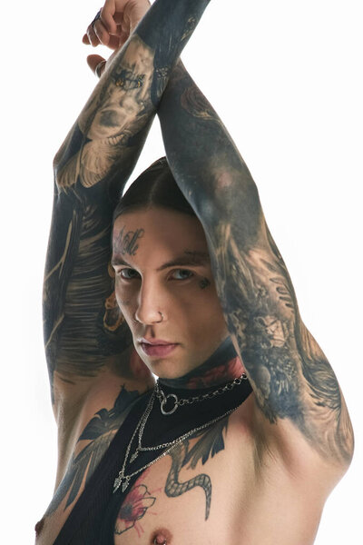 Молодой стильный мужчина с широкими татуировками и пирсингом на руках позирует в студии на сером фоне.