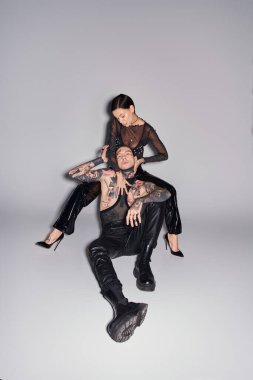 A young stylish woman is seated on top of a man in a studio, both covered in tattoos, against a grey background. clipart