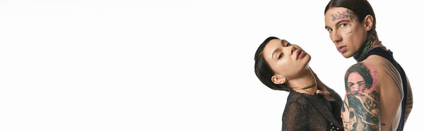 Молодой, стильный мужчина и женщина со сложными татуировками позируют в студии на сером фоне.