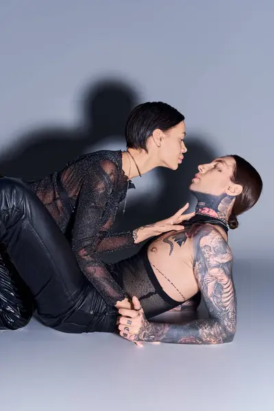 A young, stylish, and tattooed couple laying on the ground next to each other in a studio against a grey background.