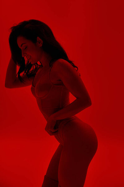 Woman in lingerie confidently stands with hands on hips in a vibrant red room.