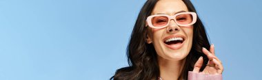 A pretty woman wearing pink sunglasses in a studio on a blue background, making a comical expression. clipart