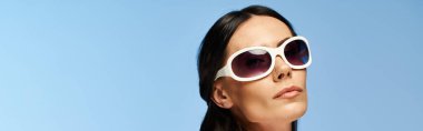 A glamorous woman wearing sunglasses looks up into the sky against a blue studio backdrop. clipart