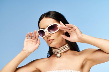 Beyaz elbiseli göz kamaştırıcı bir kadın zarif bir şekilde güneş gözlüğünü kaldırıyor ve canlı mavi bir arka plana yaz zarafeti yayıyor..