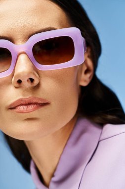 A stylish woman adorned in a purple shirt and matching sunglasses poses confidently in a studio against a vibrant blue backdrop. clipart