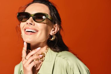 A stylish woman with sunglasses and a green shirt poses confidently against an orange backdrop in a studio setting. clipart