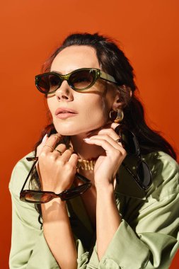 A stylish woman with sunglasses poses in a green shirt in a studio against an orange background, exuding summertime fashion vibes. clipart