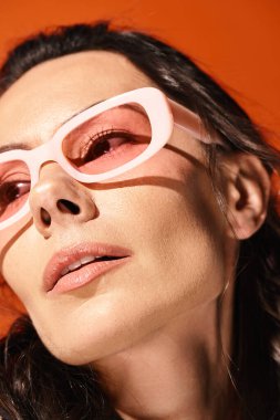 Pembe gözlüklü ve beyaz gömlekli şık bir kadın turuncu bir arka plana yaz titreşimleri yayıyor..