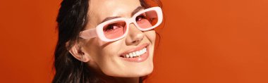 A stylish woman donning pink sunglasses smiles brightly at the camera in a vibrant studio setting against an orange background. clipart