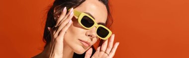 Sarı güneş gözlüğü takan şık bir kadın, ellerini yüzüne nazikçe yerleştirir. Turuncu stüdyo arka planında özgüven ve yaz modası yayar..