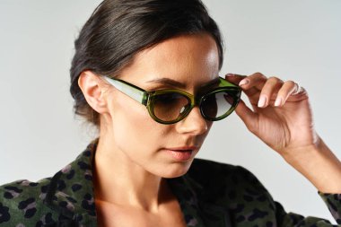 Modayı takip eden bir kadın stüdyoda gri bir arka planda spor ceket ve modaya uygun güneş gözlüğü takıyor..