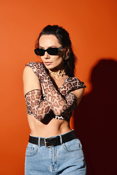 A fashionable woman poses in leopard print top and sunglasses on an orange background, exuding summertime chic.