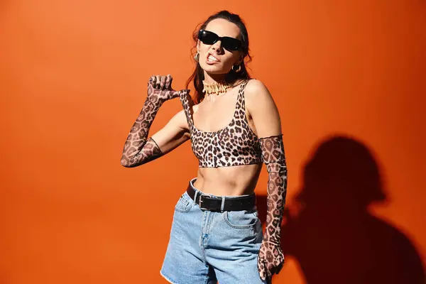 stock image A stylish woman in sunglasses stands confidently in a studio, wearing a leopard print top and denim shorts against an orange background.
