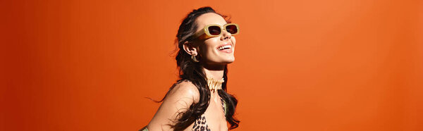 A stylish woman in a leopard print dress and sunglasses poses confidently in a studio against an orange background.