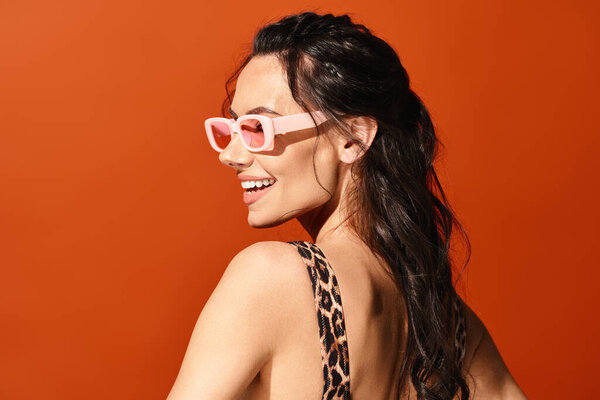 A fashionable woman exudes summertime vibes in a leopard print top, accessorized with pink sunglasses, against an orange background.