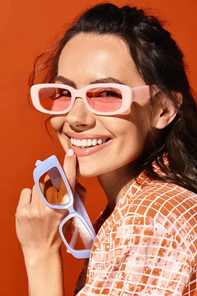 ピンクのサングラスが付いているスタイリッシュな女性は 彼女がオレンジ色の背景に微笑むように夏のバイブを露出します — ストック写真