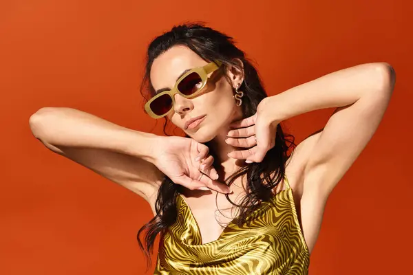 A stylish woman shines in a gold dress and trendy sunglasses against a vibrant orange backdrop.