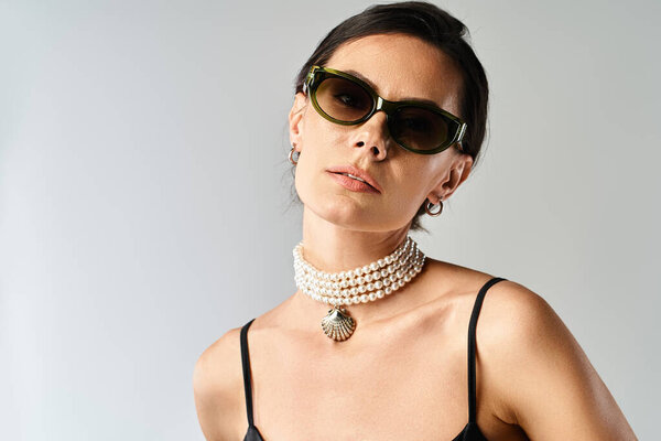 A stylish woman poses confidently wearing sunglasses and a statement necklace in a studio on a grey background.