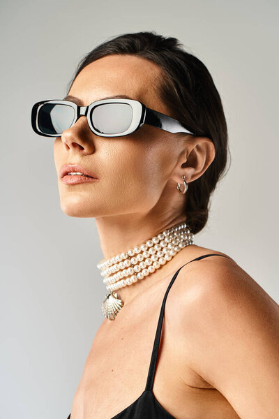 A stylish woman adorns a pair of sunglasses and pearls, exuding elegance and sophistication against a grey backdrop.