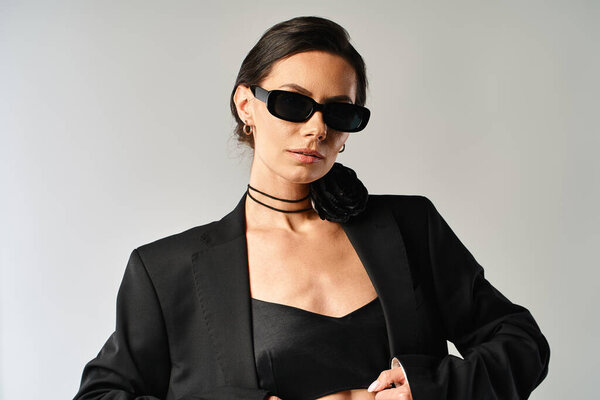 A stylish woman in a black suit and sunglasses exudes confidence and mystery in a studio with a grey background.