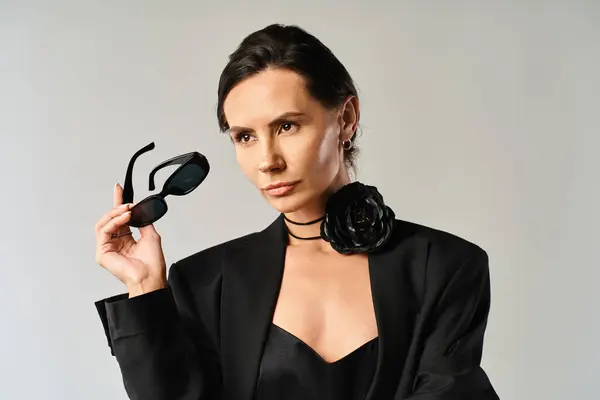 stock image A fashionable woman in a sleek black suit confidently holds up a pair of chic sunglasses, exuding a sense of sophistication and style.