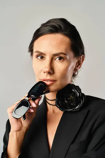 黒いスーツのスタイリッシュな女性は グレーの背景に対してスタジオで優雅さと洗練を示すサングラスを保持しています — ストック写真