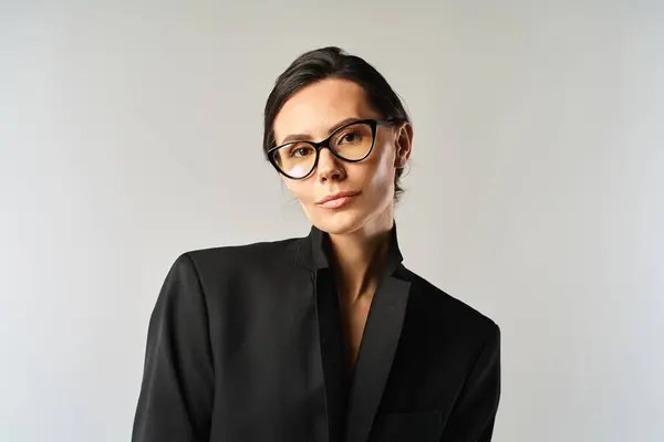 stock image A stylish woman wearing glasses and a black jacket poses in a studio against a grey background.
