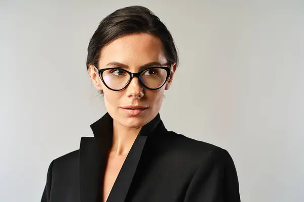 スタイリッシュな女性は 灰色のスタジオの背景に対して黒いジャケットとメガネで洗練されています — ストック写真