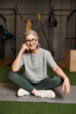 joyous mature woman in comfy sportswear sitting with crossed legs in gym and smiling at camera clipart