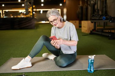 Spor kıyafetleri içinde güzel, pozitif bir kadın spor salonunda oturuyor ve akıllı telefonuna bakıyor.