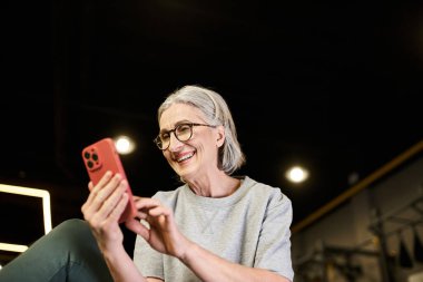 Spor kıyafetleri içinde gözlüklü neşeli olgun bir kadın spor salonunda akıllı telefonuna bakıyor.