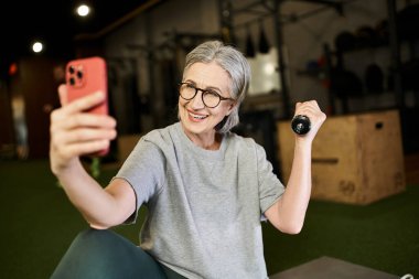 merry pretty mature woman in gray t shirt with glasses taking selfies while holding dumbbells clipart