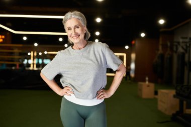 beautiful mature positive woman in cozy sportswear posing with arms akimbo and smiling at camera clipart