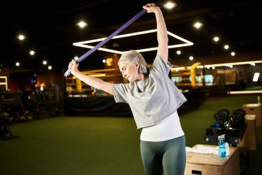 joyful senior sportswoman in comfy attire exercising actively with fitness expander while in gym clipart