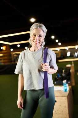 joyous senior woman in sportswear posing actively with fitness expander and smiling at camera clipart