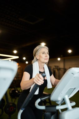 Güzel atletik olgun bir kadın spor salonunda omuzlarında havluyla antrenman yapıyor.
