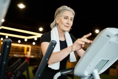 Atletik son sınıf öğrencisi, gri saçlı, omuzlarında havlusu olan, çapraz antrenör üzerinde egzersiz yapan neşeli bir kadın.