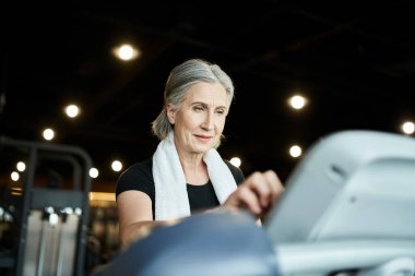 Keyifli, olgun, rahat tişörtlü, omuzlarında havluyla koşu bandında antrenman yapan spor kadını.