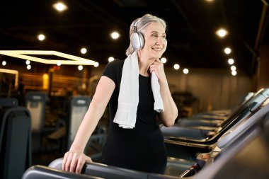 Gri saçlı, havlusu olan neşeli, olgun bir bayan müzik dinliyor ve koşu bandında egzersiz yapıyor.