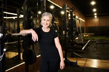 Spor kıyafetleri içinde neşeli, çekici bir kadın spor salonunda poz veriyor ve kameraya mutlu bir şekilde gülümsüyor.