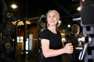 Neşeli, gri saçlı, spor kıyafetli olgun bir kadın spor salonundayken ağırlık disklerinin yanına bakıyor.