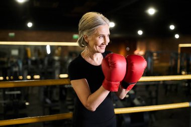 attractive jolly senior woman in sportswear with gray hair and boxing gloves posing on ring in gym clipart